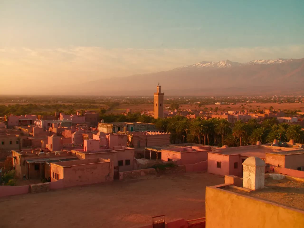 A Moroccan town at golden hour — pink earth houses, a minaret, palm groves and the snow-capped Atlas beyond